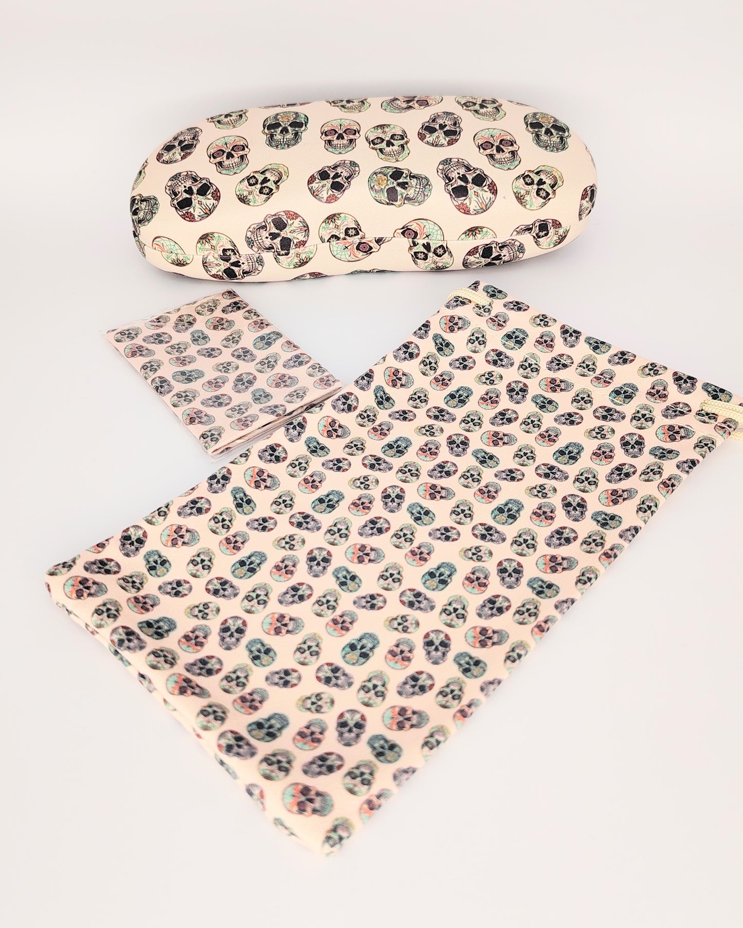 pack para gafas formado por estuche de cremallera, funda de tela blanda y gamuza limpia gafas, todo ello color beige de fondo en estampado de calaveras multicolor