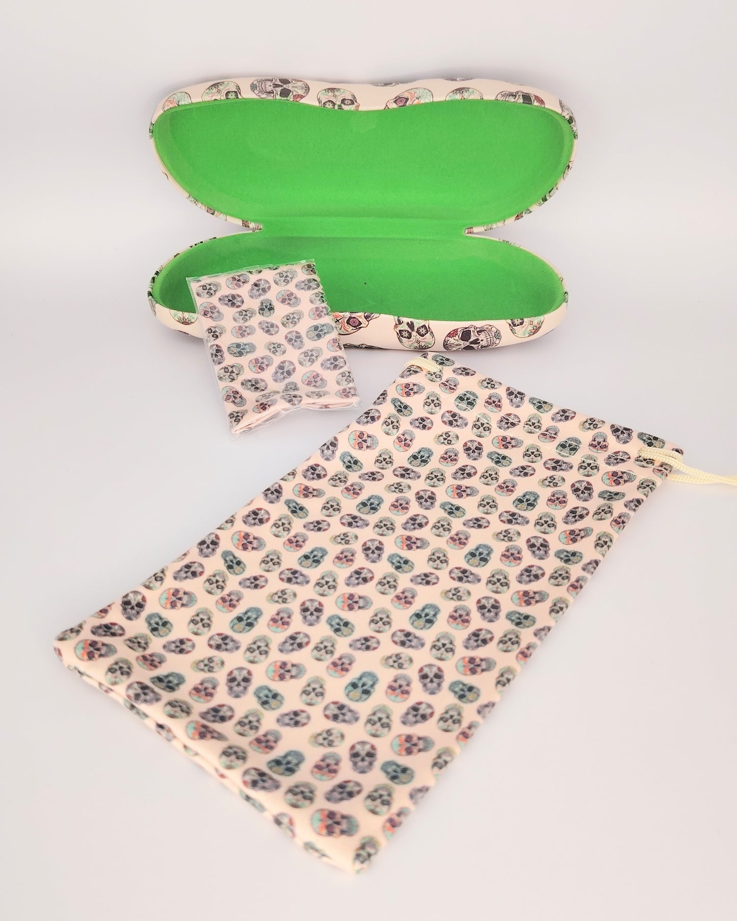 pack para gafas formado por estuche rígido, funda de tela blanda y gamuza limpia gafas, todo ello color beige de fondo en estampado de calaveras multicolor
